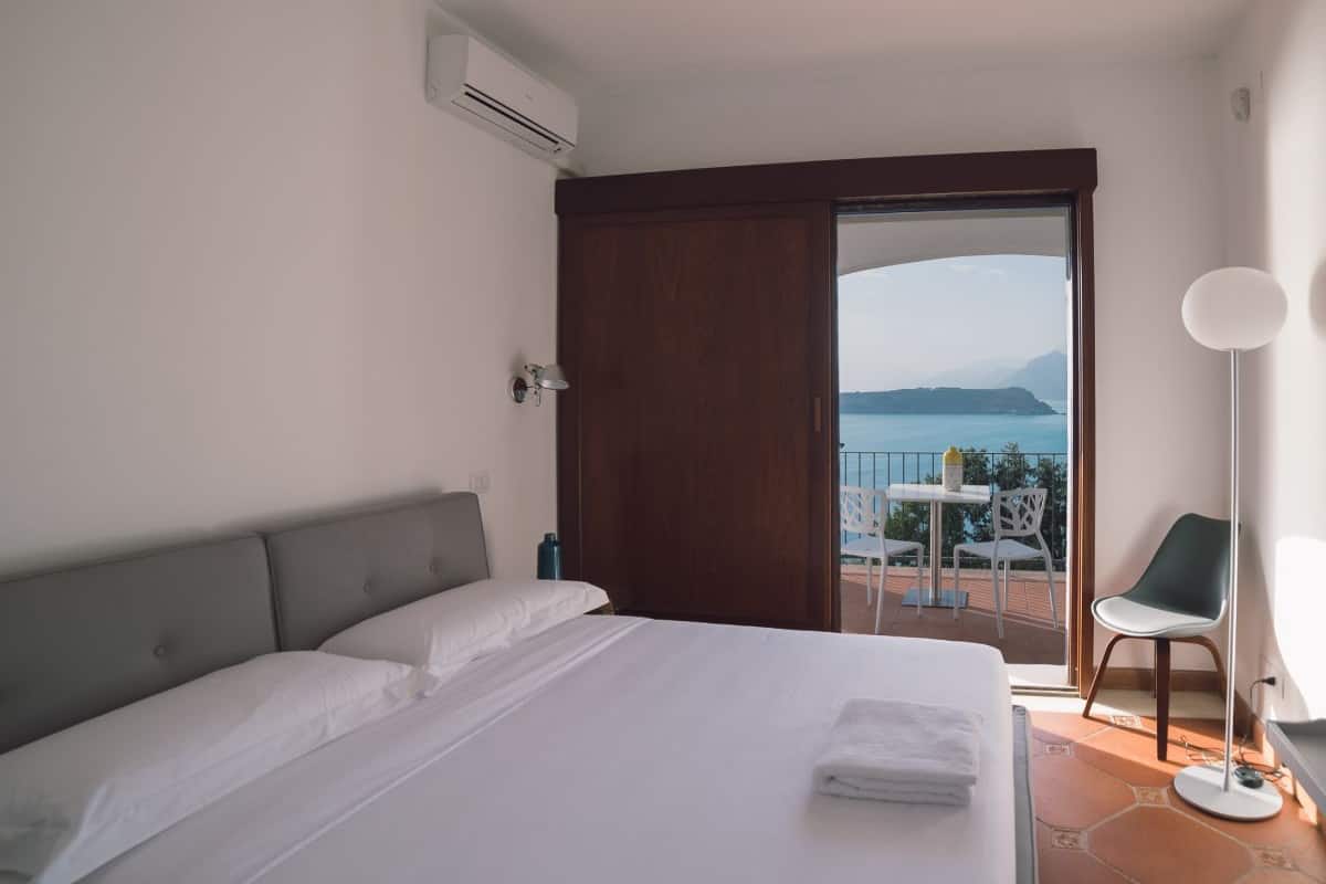 Quarto de hotel com cama, vista para o mar e varanda com cadeiras.