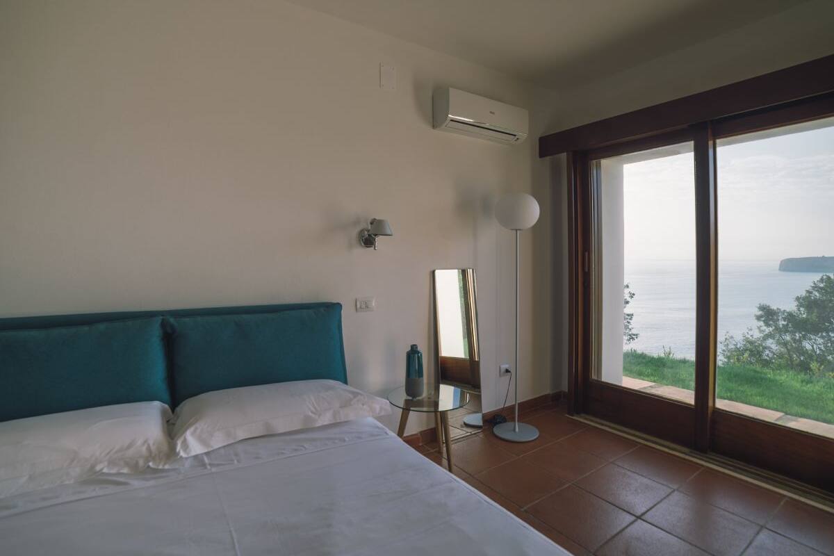 Quarto de hotel com uma cama grande, uma luminária de pé, um espelho e vista para o mar por uma janela ampla.