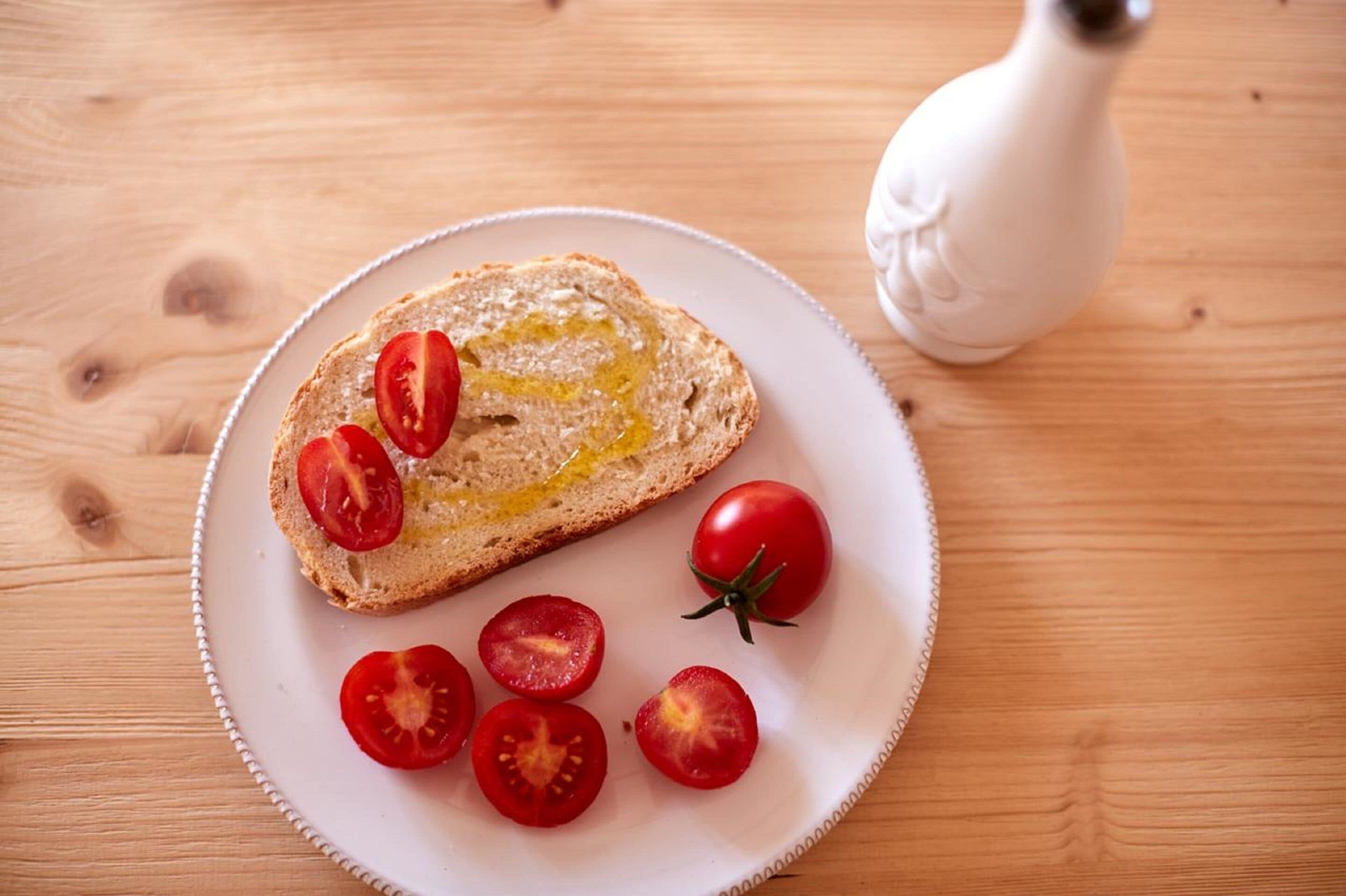 A plate with a slice of bread topped with olive oil and tomato slices, accompanied by whole and halved tomatoes on a wooden table.