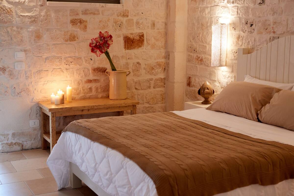 Une chambre confortable avec un mur en pierre, une table de chevet en bois ornée de bougies, un vase rempli de fleurs et un lit recouvert d’une couverture marron.