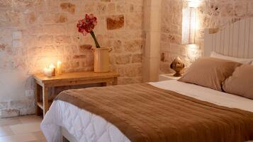 A cozy bedroom with a stone wall, a wooden nightstand with candles, a vase with flowers, and a bed with a brown blanket.