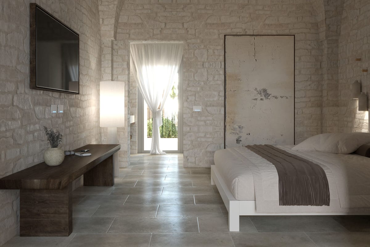 Une chambre avec un mur en pierre, un lit, un banc, une télévision et une fenêtre avec des rideaux.