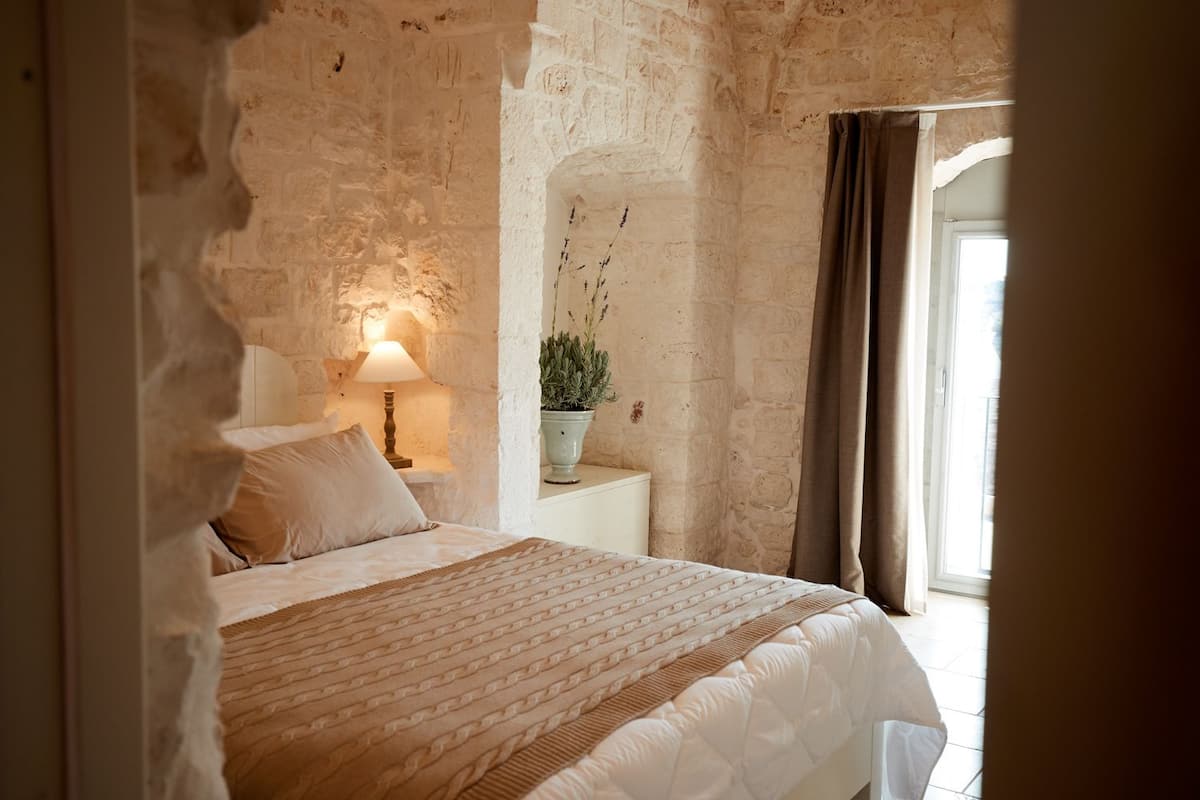 Une chambre avec un mur en briques, un lit recouvert d’une couvre-lit à motifs, une lampe de chevet et une plante en pot.