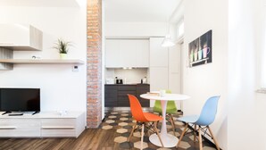 A compact living space with a TV, a small dining area with colorful chairs, a kitchen with white cabinets, and a brick accent wall. - Italianway   - Ciaia (Milan)