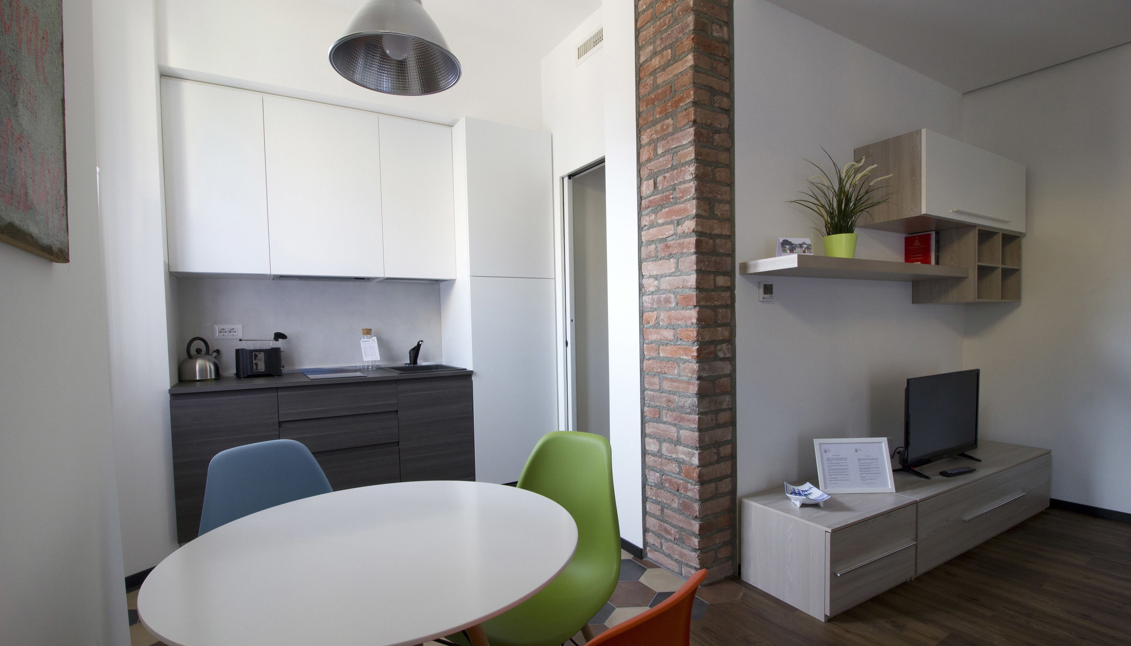 A modern kitchen with white cabinets, a sink, and a kettle. There is a dining area with a round table and two chairs. A TV is on a wooden stand, and a plant is on a shelf.