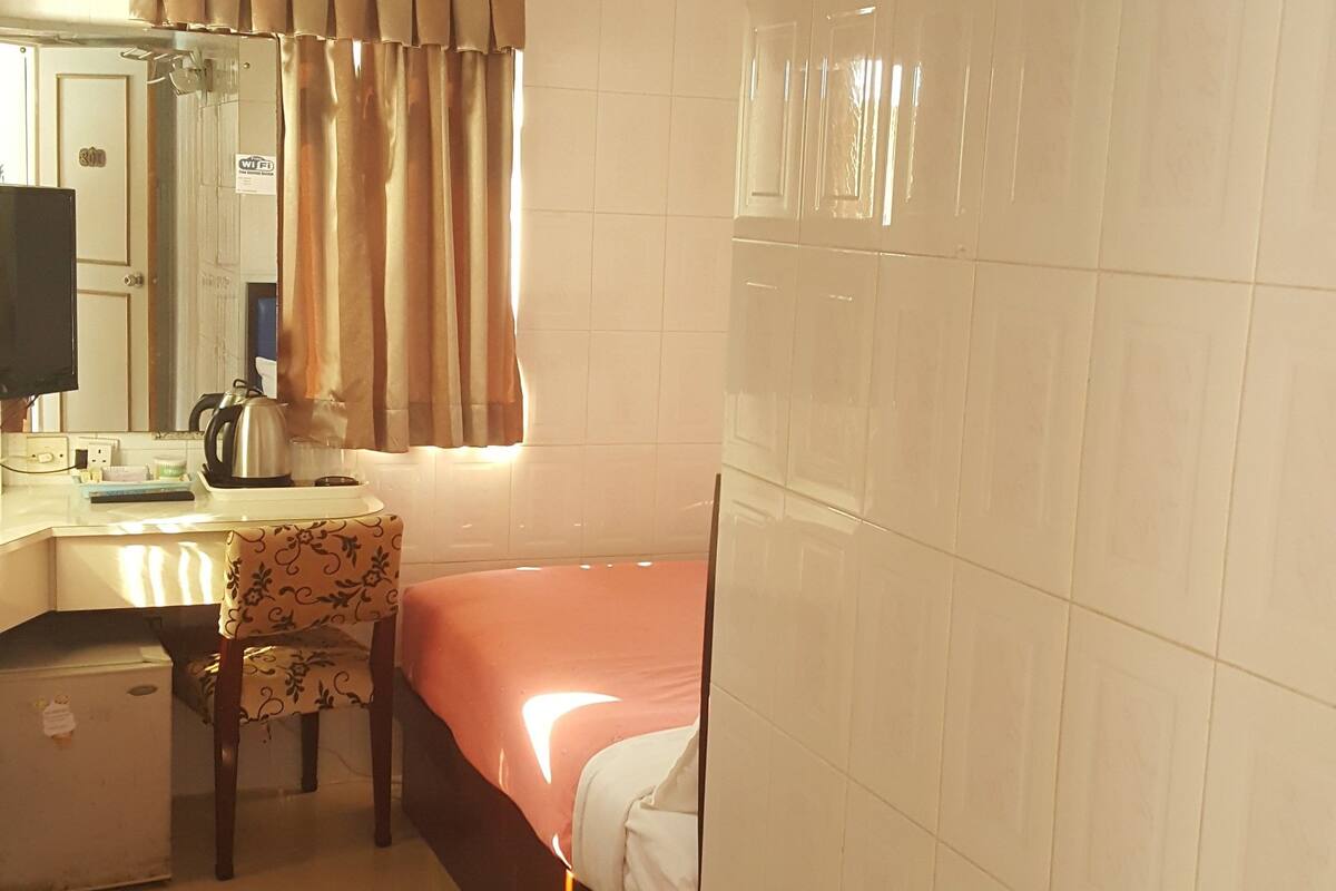Una habitación de hotel con una cama, un escritorio con hervidor, una silla, un cesto de basura y una ventana con cortinas.