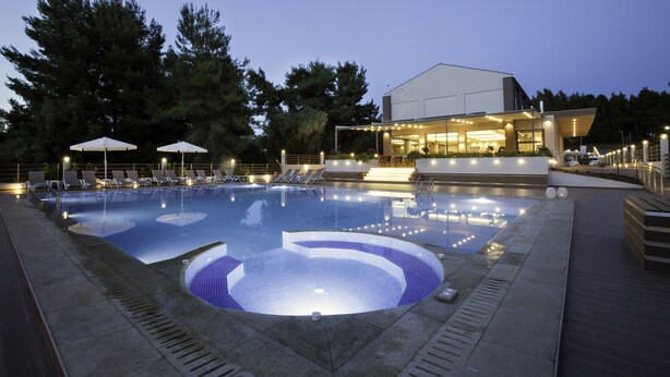 A swimming pool with lounge chairs and umbrellas, surrounded by trees and a building with illuminated windows. - Hotel Simeon (Polygyros)