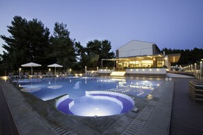 Une piscine entourée d’arbres, avec des chaises longues et des parasols, et un bâtiment dont les fenêtres sont illuminées.