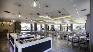 A spacious cafeteria with multiple dining tables and chairs, a buffet counter with various food items, and pendant lighting. - Hotel Simeon (Polygyros)