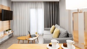 A modern hotel room with a flat-screen TV, a laptop on a small table, a sofa with cushions, and a dining table with two cups and a slice of cake. - Hotel Simeon (Polygyros)
