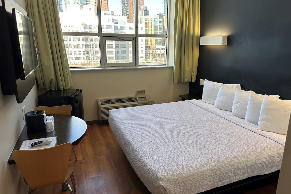 A hotel room with a large bed, a desk, a chair, a TV, and a window with a city view.