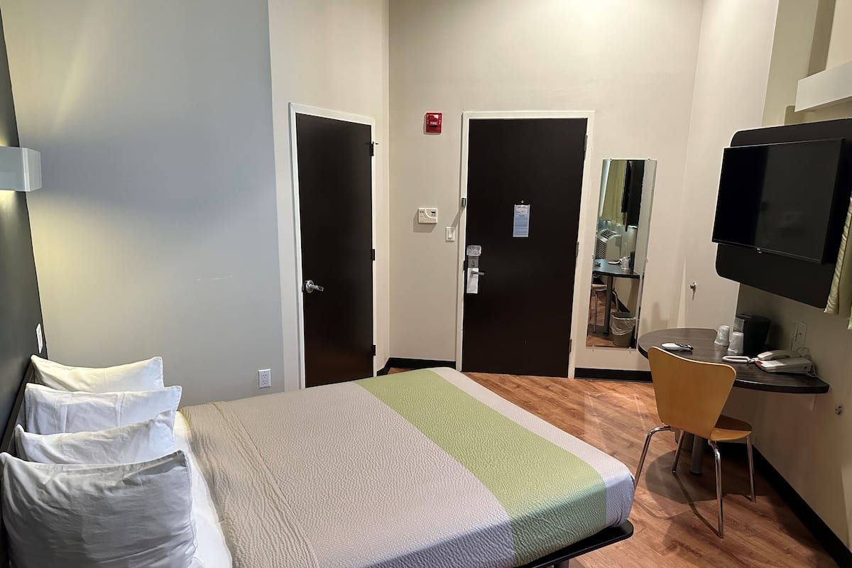 A hotel room with a bed, a chair, a desk with a computer, a TV, and a door with a fire alarm.