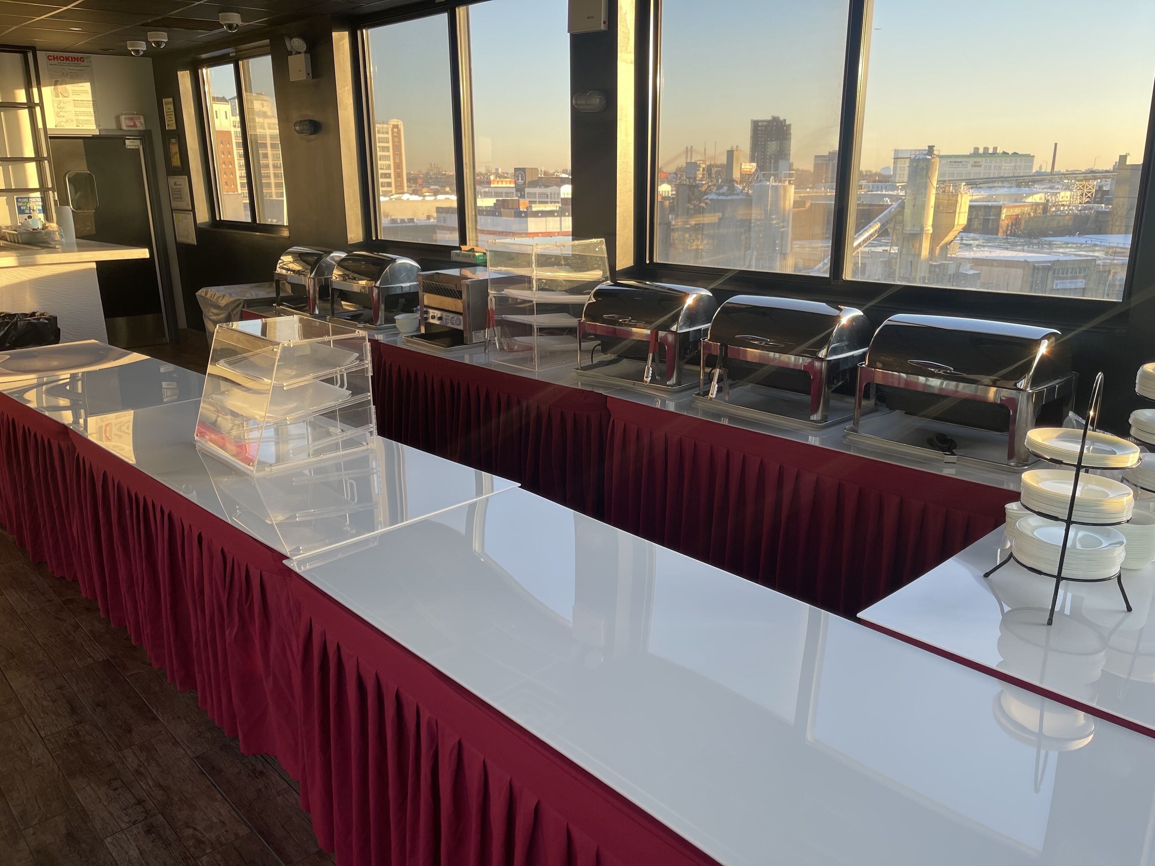 A buffet setup with multiple self-service stations, a red tablecloth, and a city view through large windows.