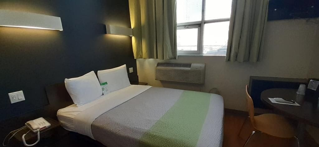 A hotel room with a bed, a desk, a chair, a window with curtains, an air conditioning unit, and a phone on the nightstand.