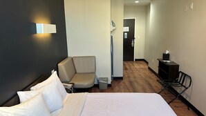 A hotel room with a large bed, a seating area with a chair, a desk with a lamp, and a trash bin.