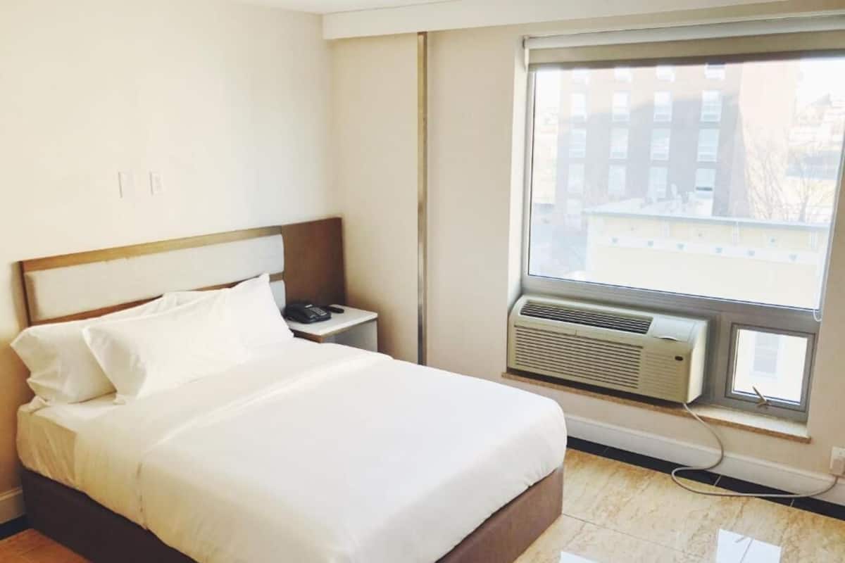 A hotel room with a bed, a nightstand, an air conditioning unit, and a window with a view of buildings.