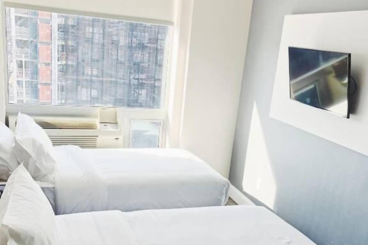 A hotel room with two beds, a large window, an air conditioning unit, and a mounted TV.