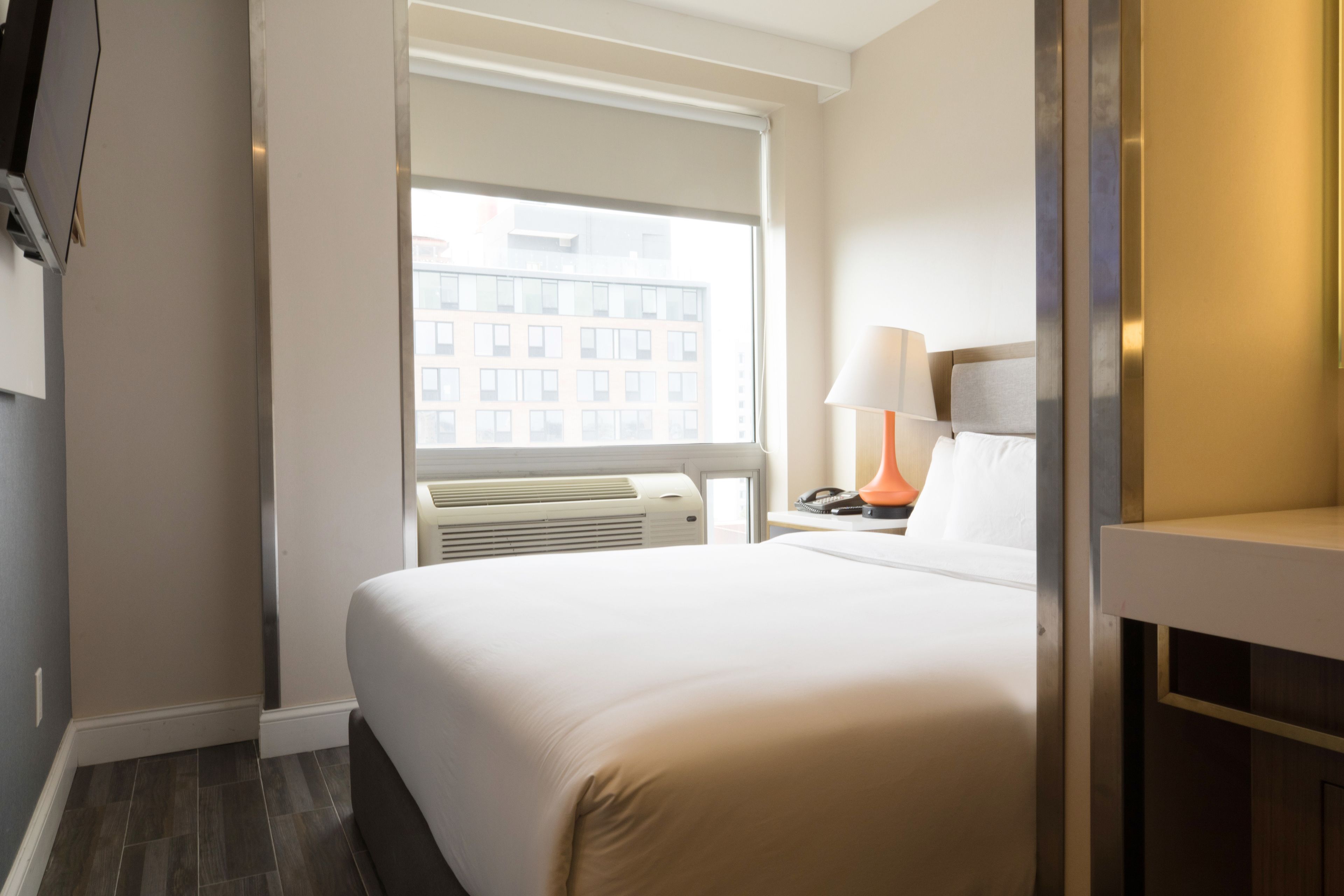 A hotel room with a large bed, a window with blinds, a bedside table with a lamp, and a wall-mounted air conditioning unit.