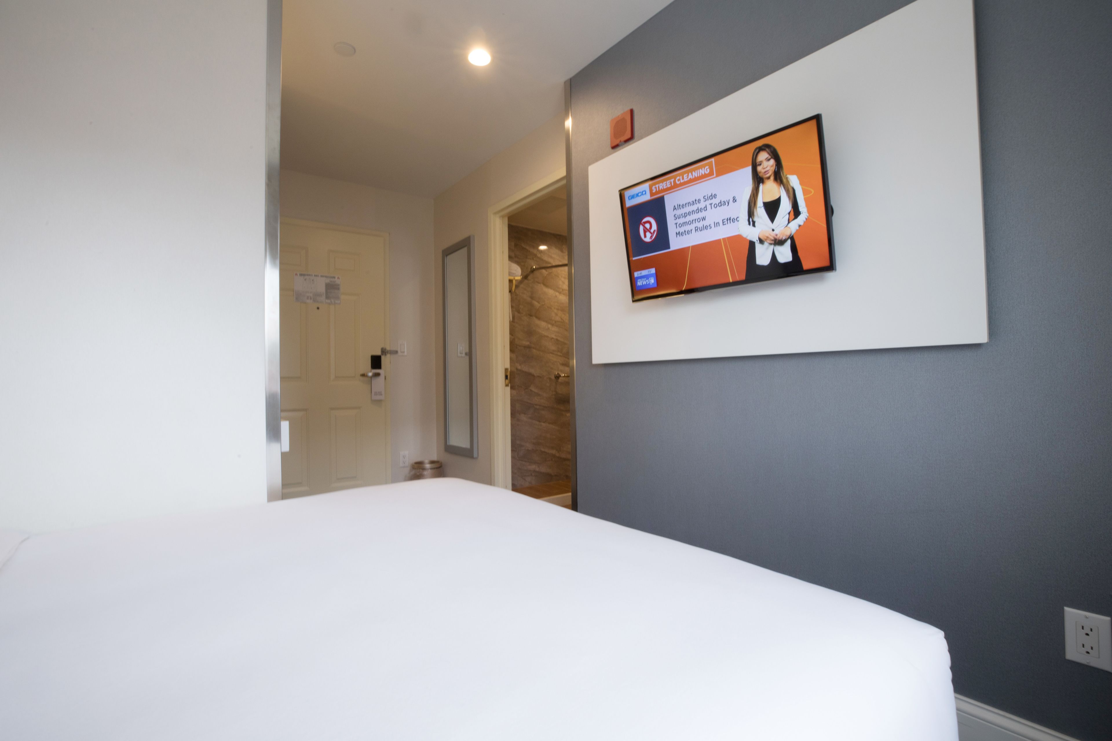 A hotel room with a bed, a television mounted on the wall displaying a news program, and a door leading to another room.