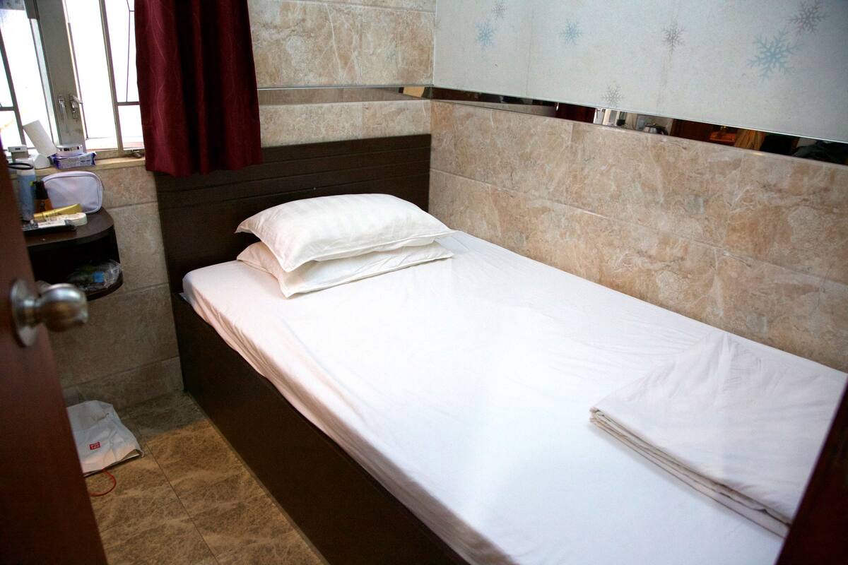A single bed with white linens in a room with beige tiled walls and a dark headboard.