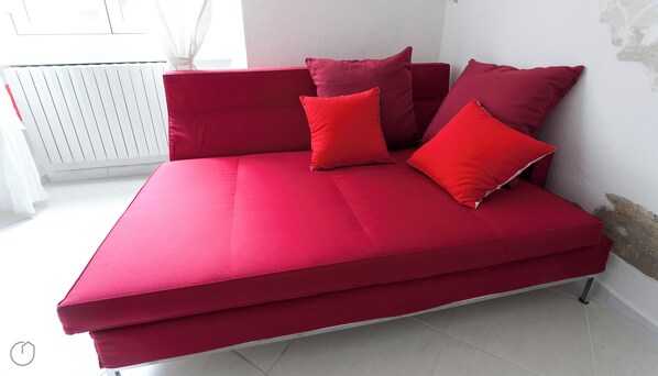 Un canapé-lit rouge avec des coussins assortis, dans une pièce aux murs blancs et dotée d’un radiateur.