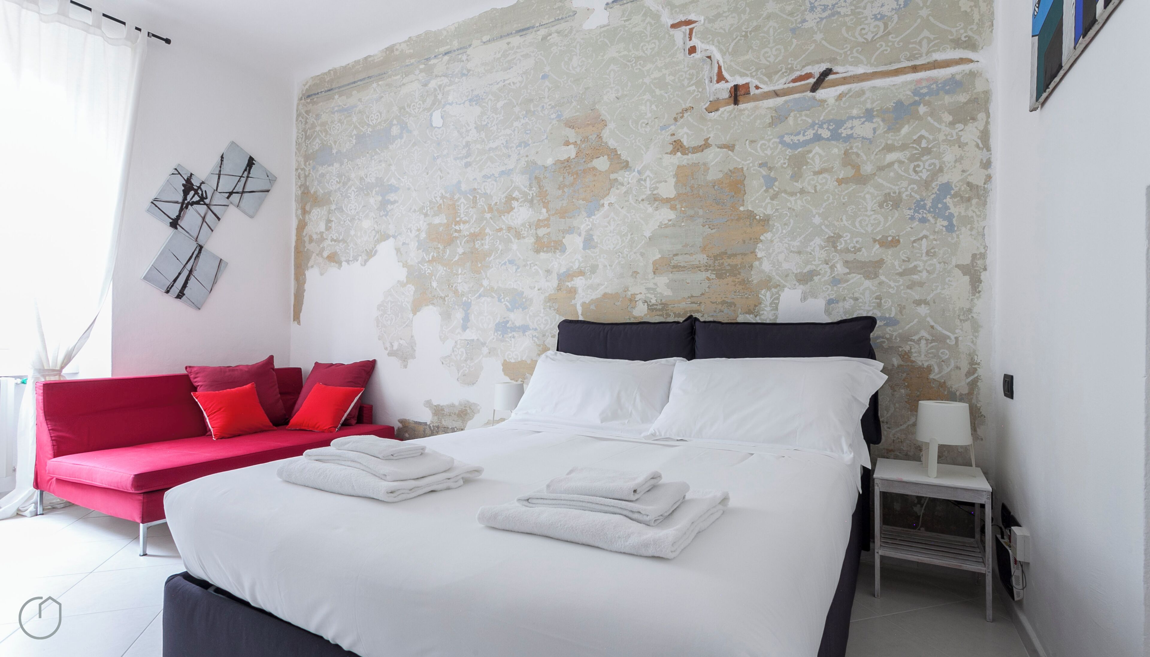 A bedroom with a bed, a red sofa, and a wall with peeling paint.