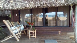 A wooden deck with a thatched roof, a wooden chair, a small table with a vase of flowers, and a view of the beach and ocean through large windows.