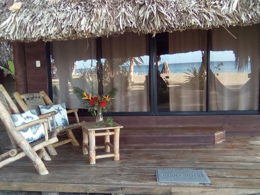 En træterrasse med stråtæthed, en træstol, et lille bord med en vase blomster og udsigt til stranden og havet gennem store vinduer.
