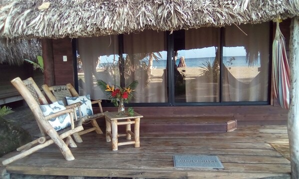 A wooden deck with a thatched roof, a wooden chair, a small table with a vase of flowers, and a view of the beach and ocean through large windows. - Playa Paraiso en Magante (Gaspar Hernandez)