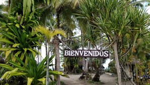 A sign reading "BIENVENIDOS" at the entrance of a tropical garden area with various plants and a dirt path.