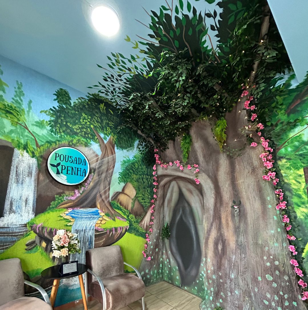 Una habitación con un mural de una escena de bosque, un cartel que dice "Pousada Penha" y una pequeña mesa con un florero de flores.