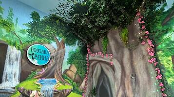 Una habitación con un mural de una escena de bosque, un cartel que dice "Pousada Penha" y una pequeña mesa con un florero de flores.