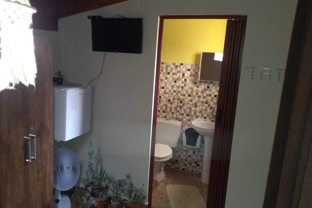 A compact bathroom with a toilet, sink, and a small fan. A refrigerator is mounted on the wall, and a flat-screen TV is visible in the background.