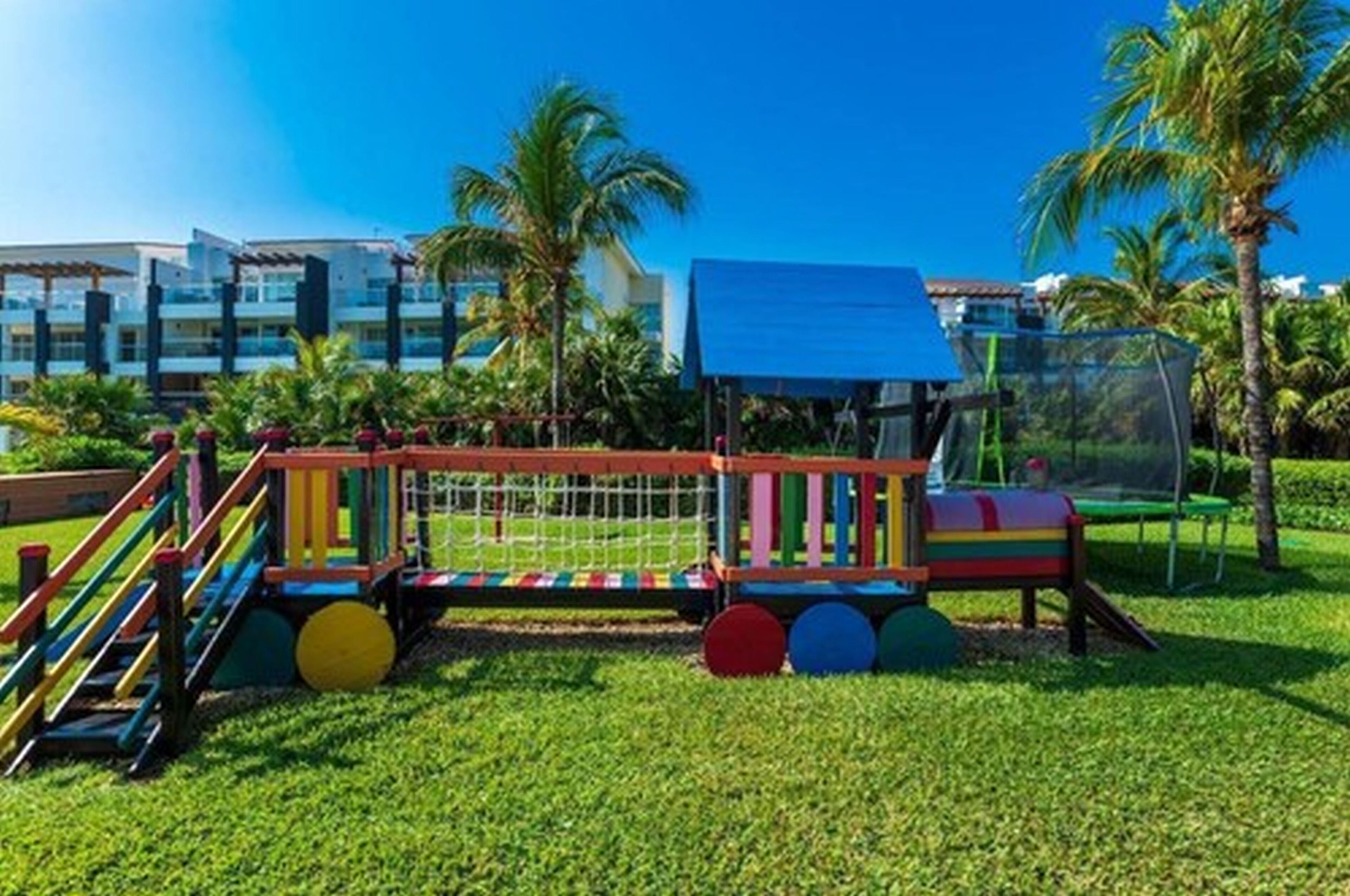 A colorful playground with a slide and climbing structures, surrounded by well-maintained grass and palm trees, with a building in the background.