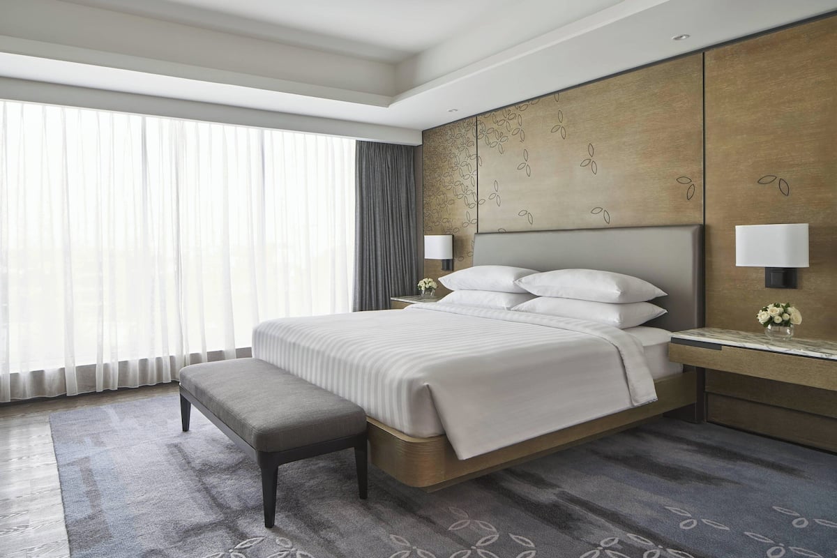 Habitación de hotel moderna con una cama grande, un banco, una mesita de noche con lámpara y un amplio ventanal con cortinas.