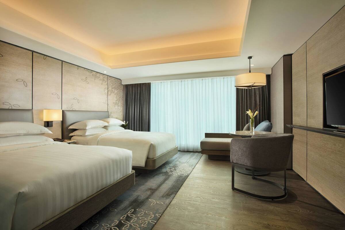 Una habitación de hotel moderna con dos camas, una zona de estar, un televisor y un amplio ventanal con cortinas.