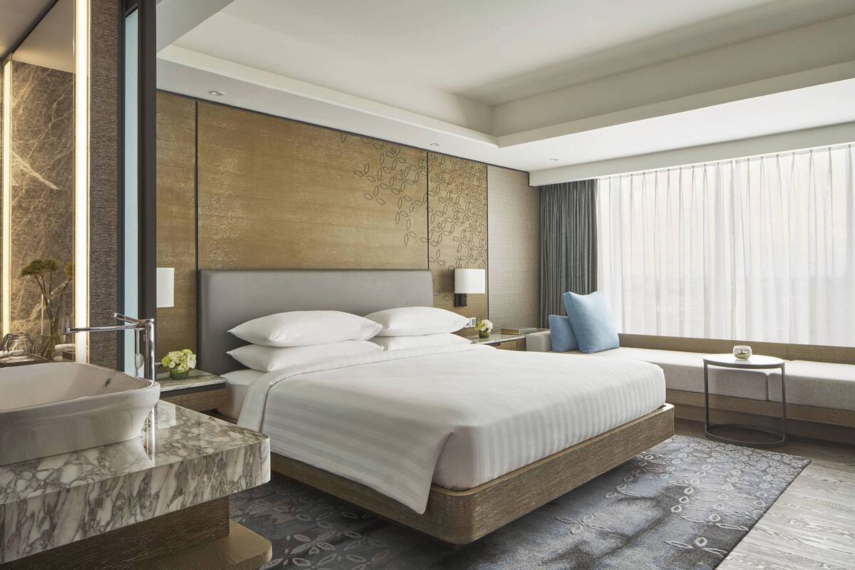Una habitación de hotel moderna con una cama grande, un lavamanos de mármol y un amplio ventanal con cortinas.