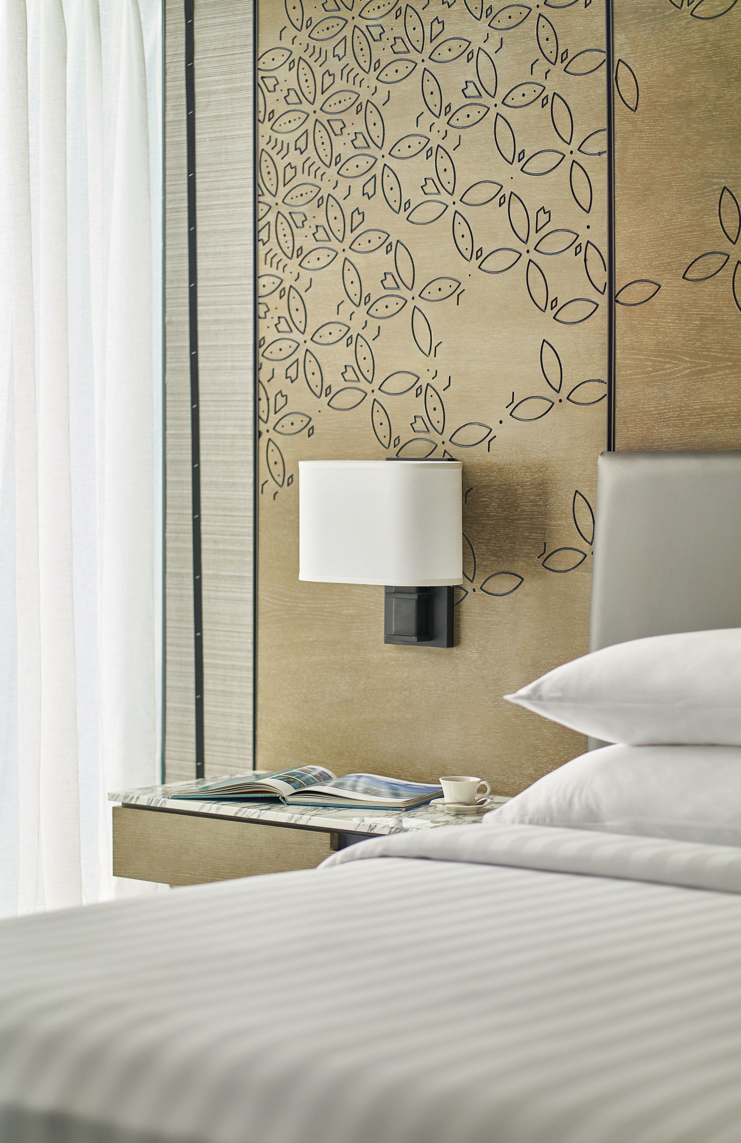 A neatly made bed with white linens, a bedside table with a lamp, and a wall with a leaf pattern.