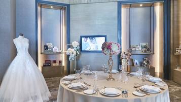 Un vestido de novia sobre un maniquí, una mesa preparada para una comida y un televisor mostrando una escena de boda.