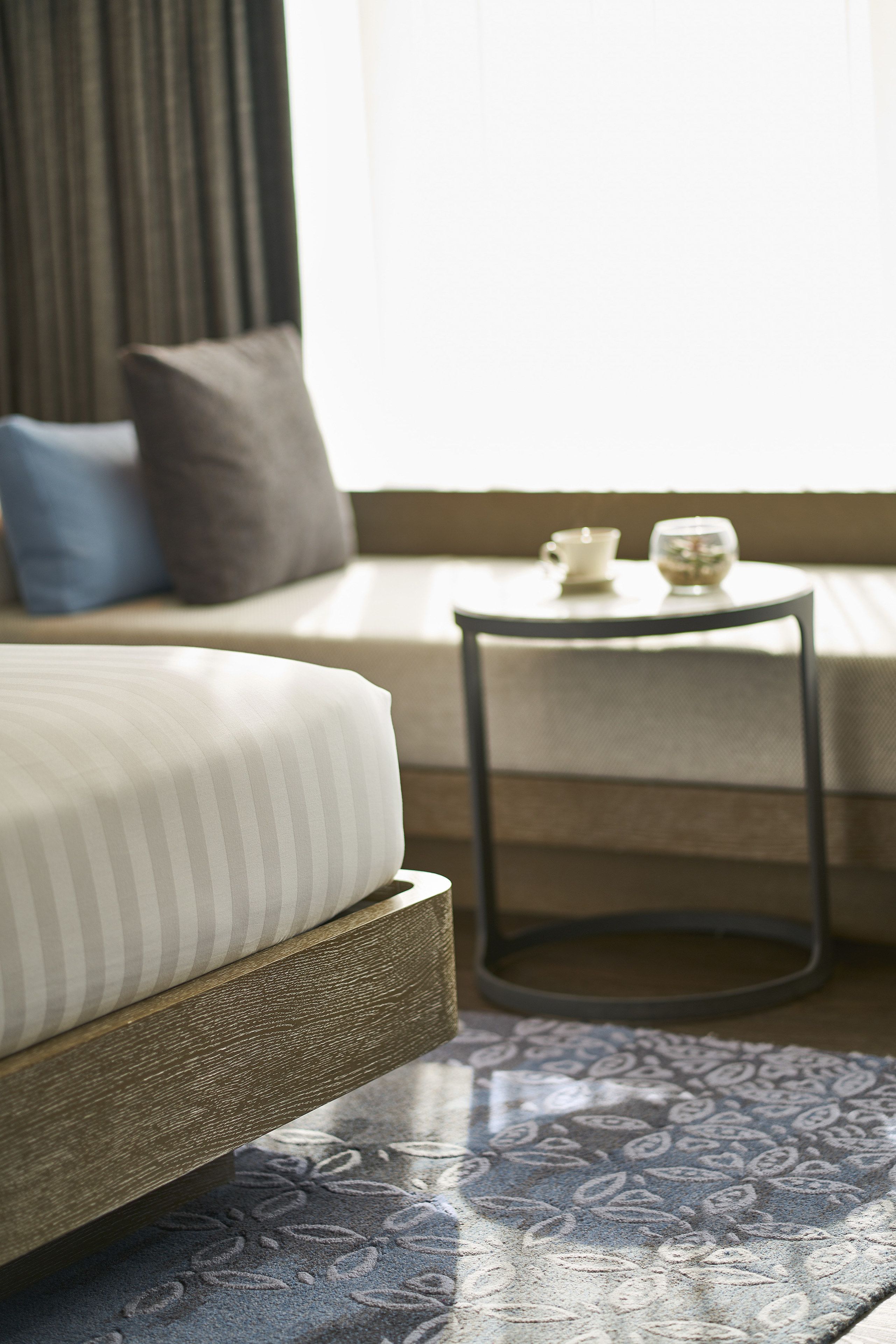 A bed with a wooden frame and a patterned rug, a small round table with a cup and glass, and a sofa with cushions.