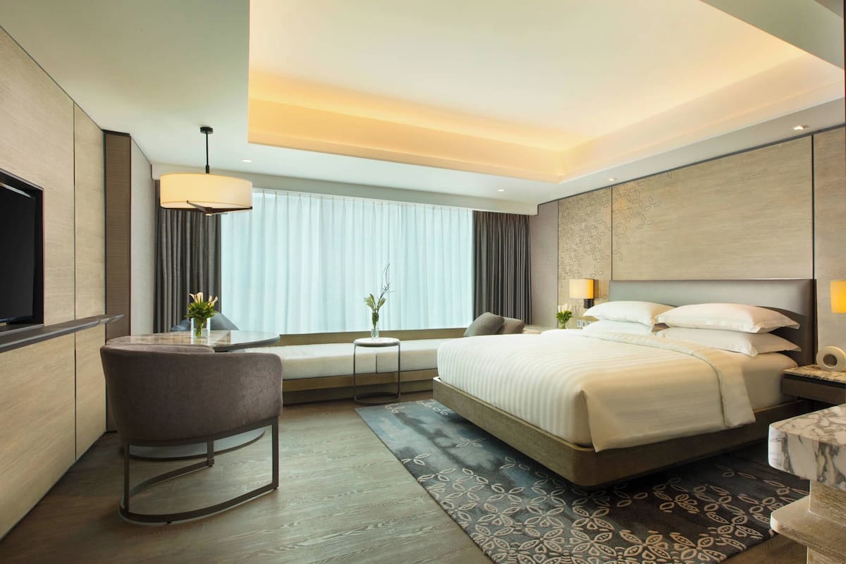Una habitación de hotel moderna con una cama grande, una zona de estar con una silla y una mesita, un televisor y un amplio ventanal con cortinas.