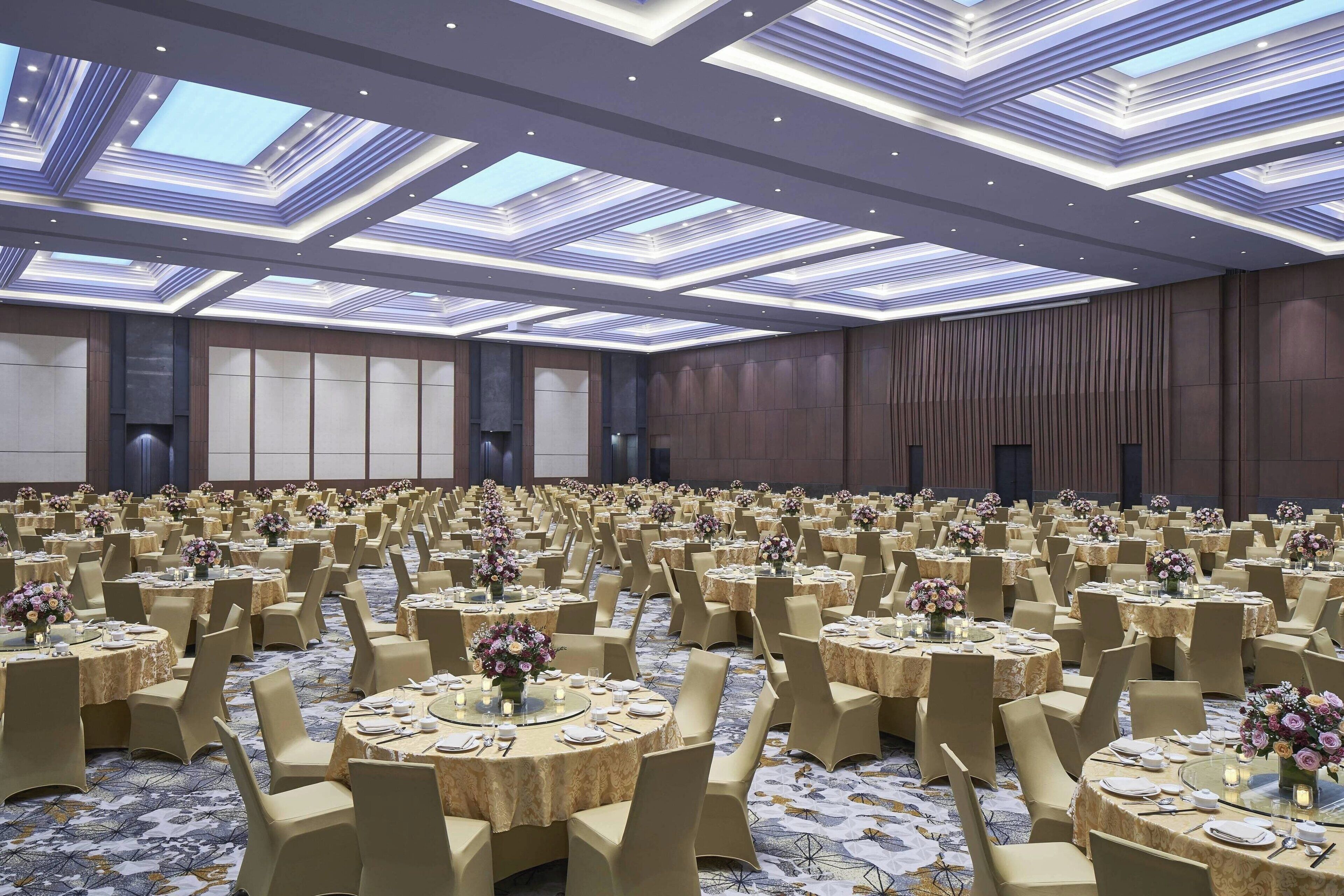 Un amplio salón de banquetes con mesas redondas, sillas color beige y centros de mesa con flores.