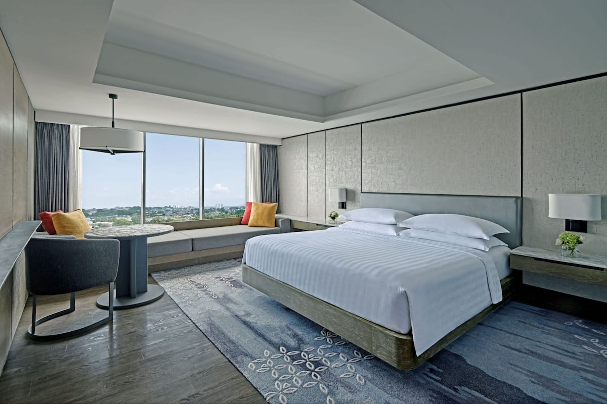 Una habitación de hotel moderna con una cama grande, un espacio de descanso con una silla y una mesita, un ventanal con vistas y una lámpara en el techo.