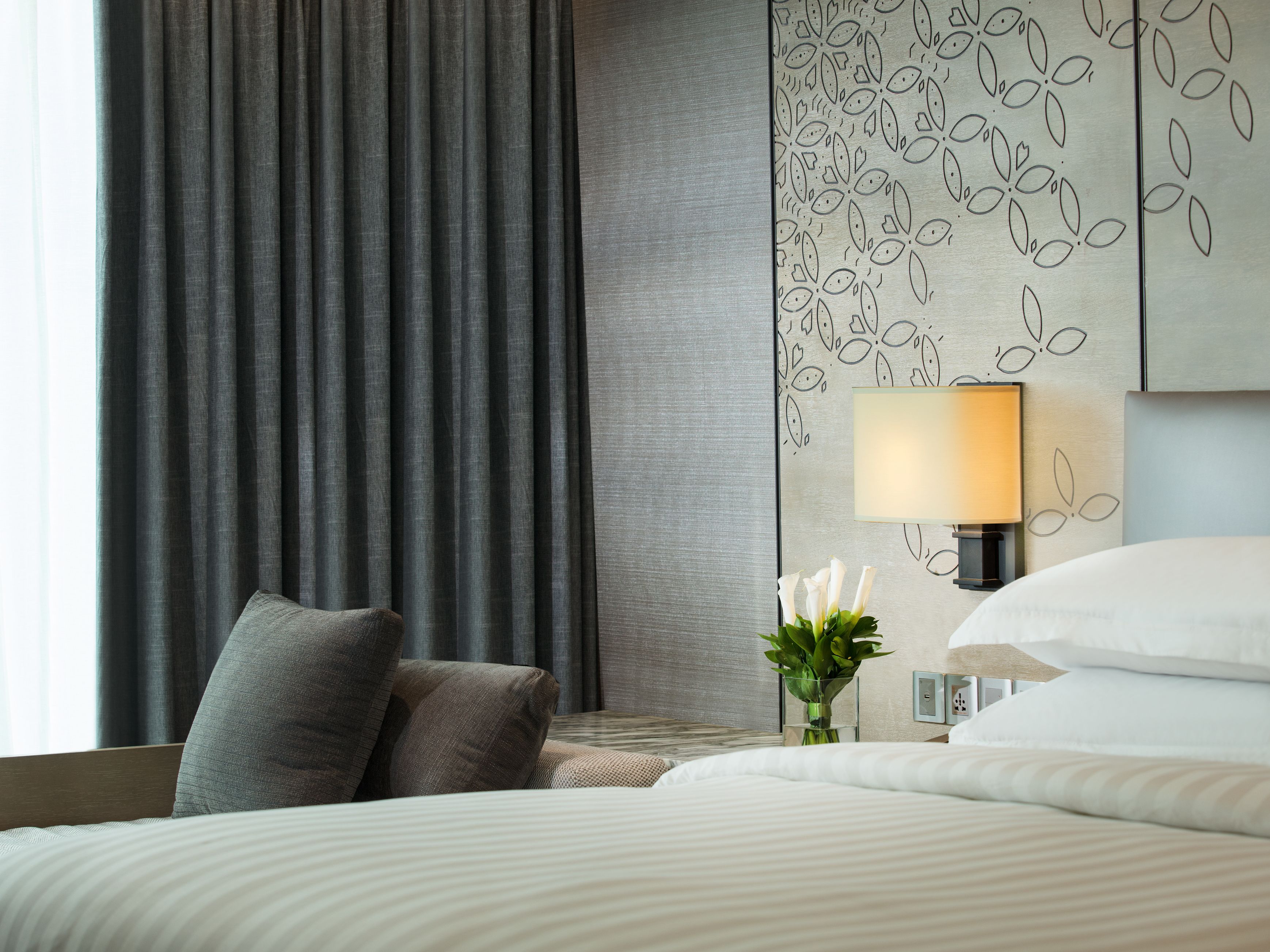 Une chambre d’hôtel avec un grand lit, une table de chevet avec une lampe, un vase de fleurs et une fenêtre avec des rideaux.