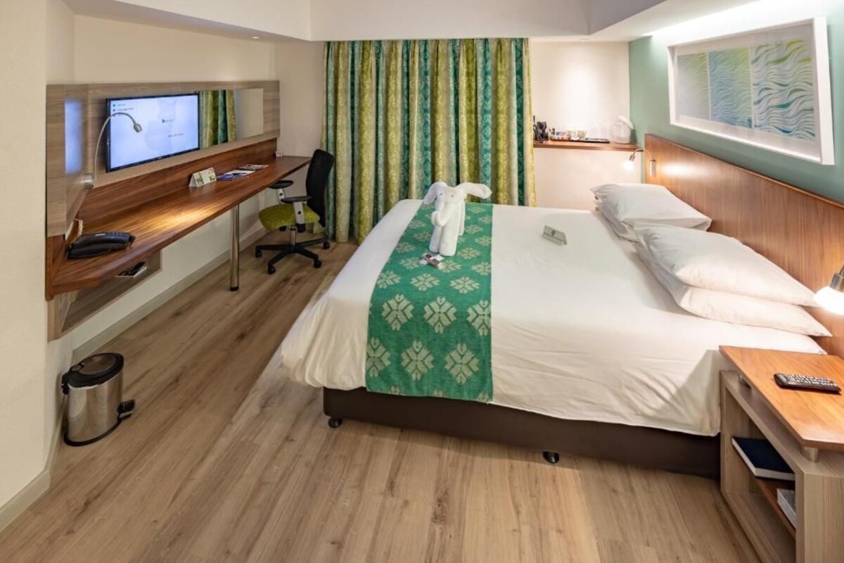 A hotel room with a bed, a desk with a computer, a trash bin, and a window with curtains.