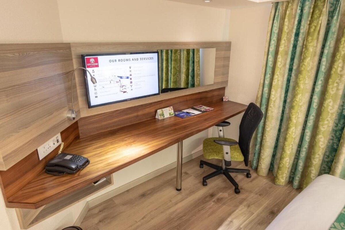 A hotel room with a wooden desk, a computer monitor displaying room and service information, a telephone, and a green patterned curtain.