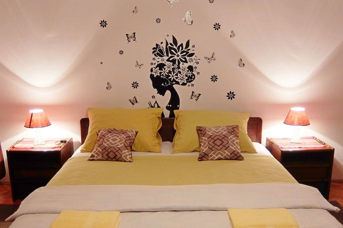 Una cama doble con dos almohadas amarillas, un mural decorativo y dos lámparas de noche.
