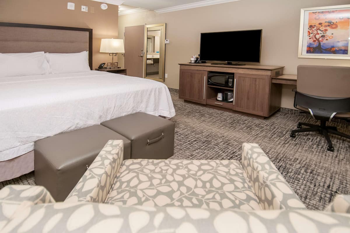 A hotel room with a large bed, a desk with a television, a chair, and a small kitchenette.