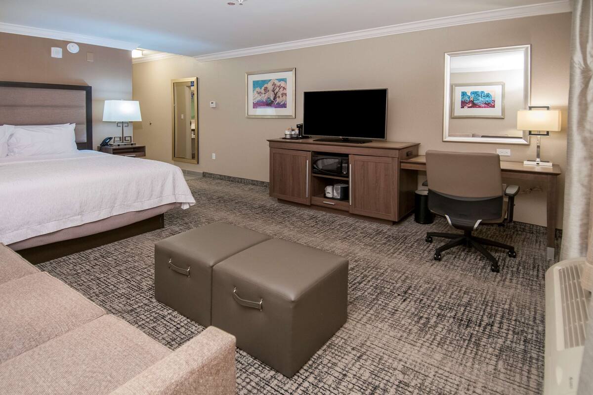 A hotel room with a bed, a desk with a computer, a TV, and a seating area with ottomans.