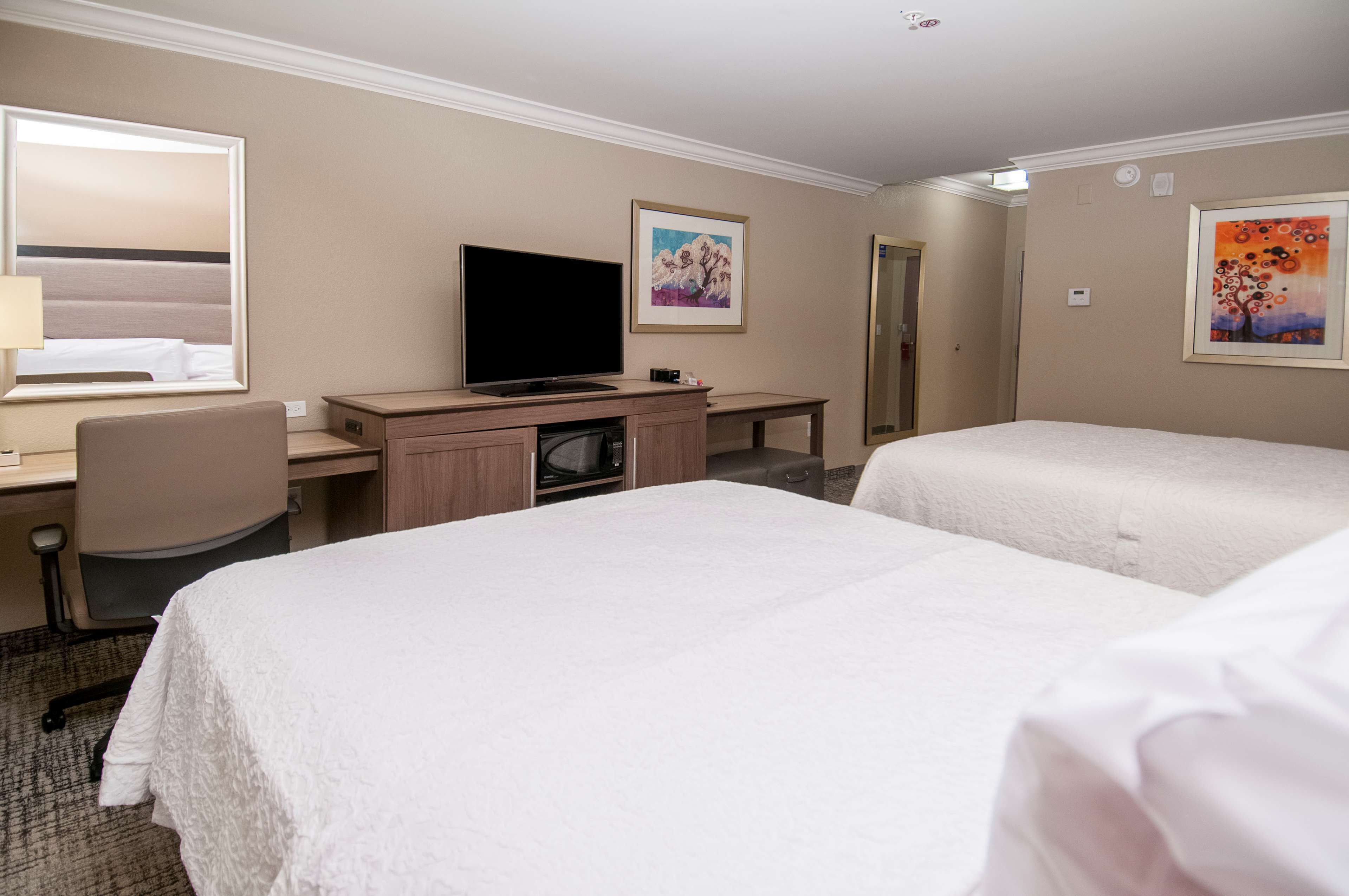 A hotel room with two beds, a desk with a television, and artwork on the walls.